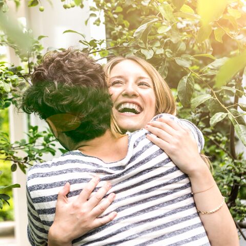 Ein lachendes Paar umarmt sich im Freien, umgeben von grünem Laub und Sonnenschein, beide tragen gestreifte Oberteile.