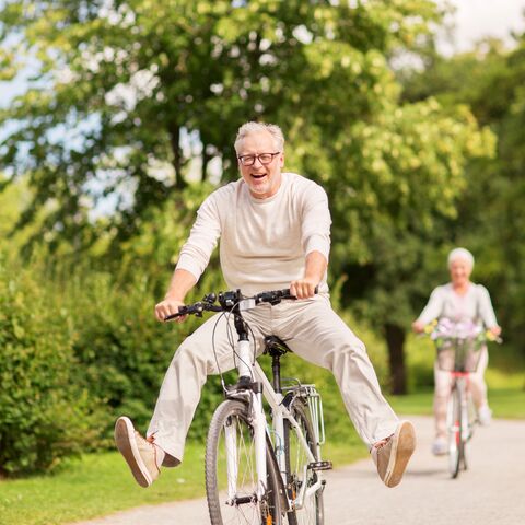 Un homme âgé fait joyeusement du vélo, les jambes surélevées, tandis qu'une femme fait également tranquillement du vélo à l'arrière-plan, entourée d'une nature verdoyante.