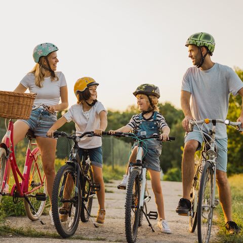 Une joyeuse famille casquée roule à vélo sur un chemin ensoleillé dans la verdure tout en discutant.