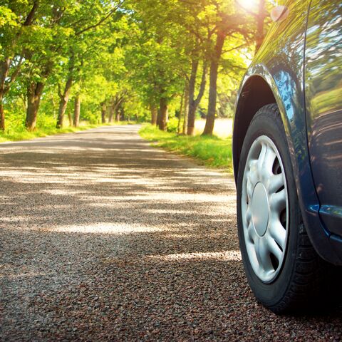 Ein blaues Auto fährt auf einer schattigen, von Bäumen gesäumten Landstrasse an einem sonnigen Tag.