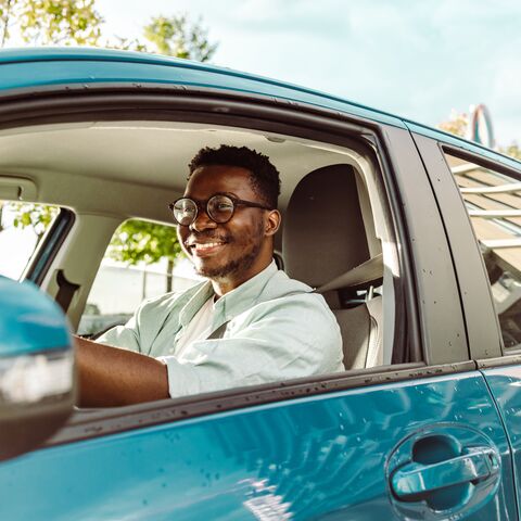 Ein lächelnder junger Mann mit Brille fährt an einem sonnigen Tag ein blaues Auto.
