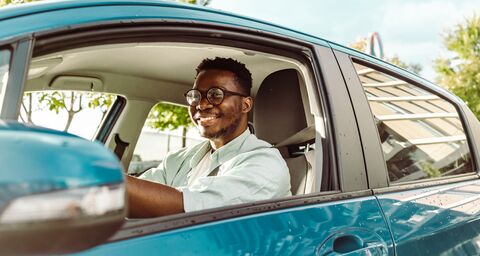 Ein lächelnder junger Mann mit Brille fährt an einem sonnigen Tag ein blaues Auto.