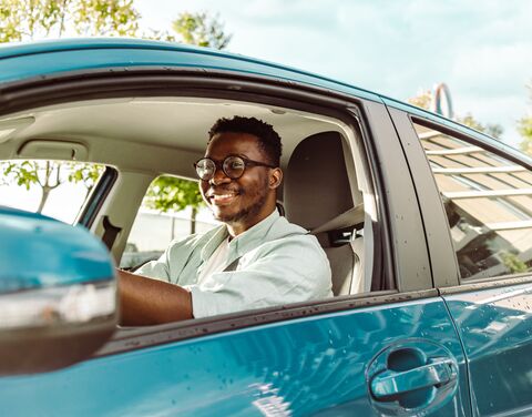 Ein lächelnder junger Mann mit Brille fährt an einem sonnigen Tag ein blaues Auto.