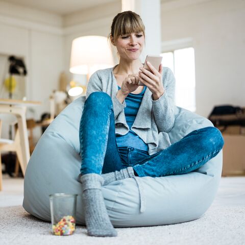 Une femme sourit, assise sur un canapé, et tape un message sur son téléphone portable.