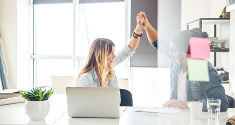 Zwei Personen in einem modernen Büro geben sich ein High-Five, während sie an einem Tisch mit Laptop und Notizen sitzen, lächelnd und motiviert.