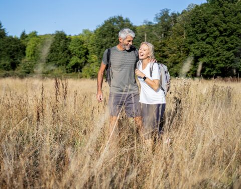 Ein älteres Paar wandert lächelnd durch eine sonnige Wiese mit hohem Gras, umgeben von Bäumen im Hintergrund.