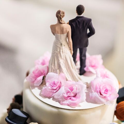 Figure di una coppia di sposi con le spalle alla telecamera, in abiti da sposa su una superficie bianca e rosa.