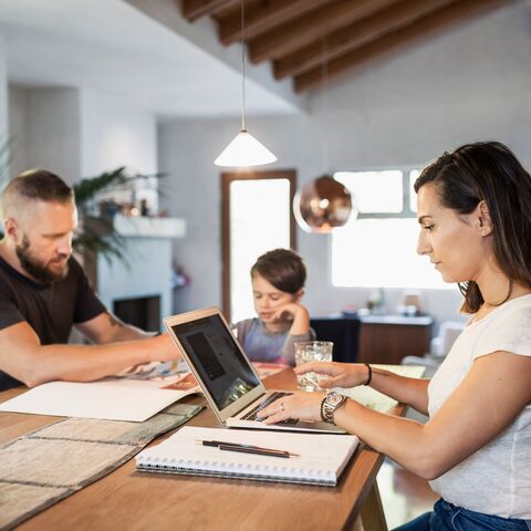 Une femme travaille sur un ordinateur portable tandis qu’un homme et un enfant sont assis à la table; en arrière-plan, on voit un salon confortable.