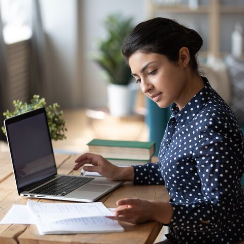 Une femme est assise à un bureau avec un ordinateur portable et étudie des documents, entourée de livres et de plantes dans une pièce lumineuse.