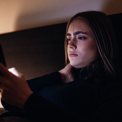 A young girl lies on her bed in a dark room and looks critically at her cell phone. 