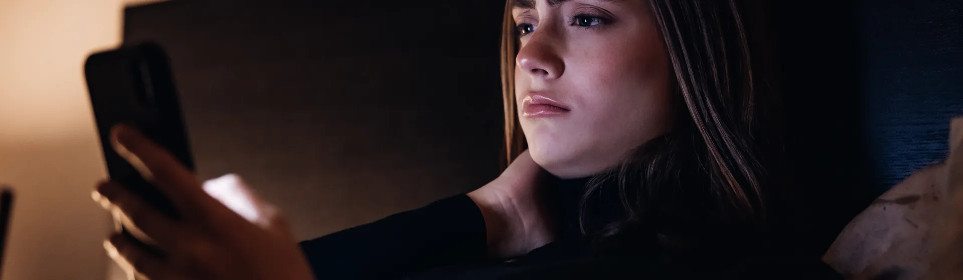 A young girl lies on her bed in a dark room and looks critically at her cell phone. 