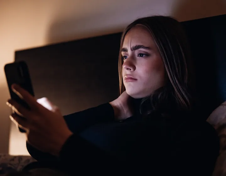Un’adolescente giace sul suo letto in una stanza buia e guarda criticamente il suo cellulare. 
