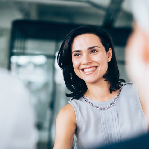 A smiling woman with shoulder-length dark hair in a gray sleeveless top chats in a modern office.