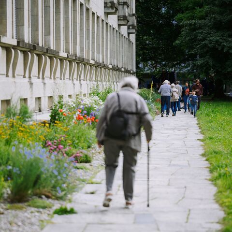 Une personne âgée marche avec une canne le long d’un chemin bordé de parterres de fleurs et d’espaces verts. D’autres personnes marchent plus loin sur le même chemin.