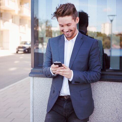 Ein junger Mann mit weissem Hemd und blauem Jacket schaut lächelnd auf sein Handy.