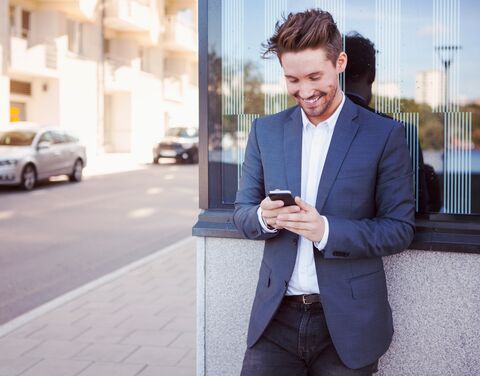 Un giovane uomo in camicia bianca e giacca blu guarda il suo cellulare sorridendo.