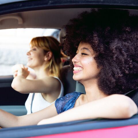 Deux femmes joyeuses sont assises dans une voiture, la conductrice rit et la passagère montre du doigt par la fenêtre, toutes deux profitent du moment.
