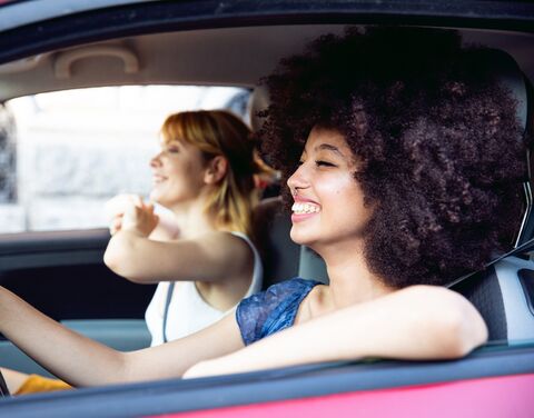 Two cheerful women are sitting in a car, the driver is laughing and the passenger is pointing out of the window, both enjoying the moment.