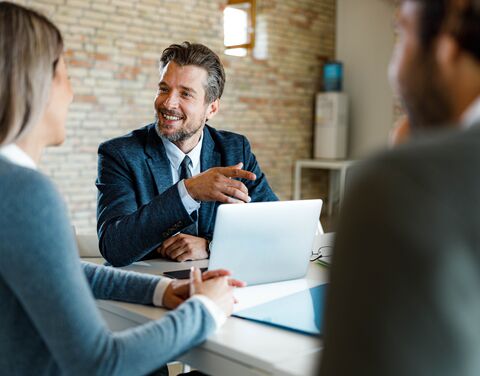 Un homme en tenue d'affaires avec un ordinateur portable en conversation avec une femme et un homme, visibles de dos, assis à une table. 