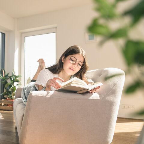 Une femme est allongée sur le canapé de son appartement et lit un livre.