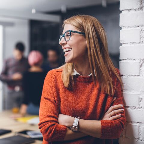 Eine junge Frau mit Brille und roten Pullover lacht fröhlich, während sie sich in einem modernen Raum mit unscharf im Hintergrund sichtbaren Menschen befindet.
