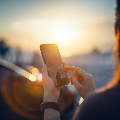 Une personne tient un smartphone à la main et y regarde un graphique financier au coucher du soleil en plein air.