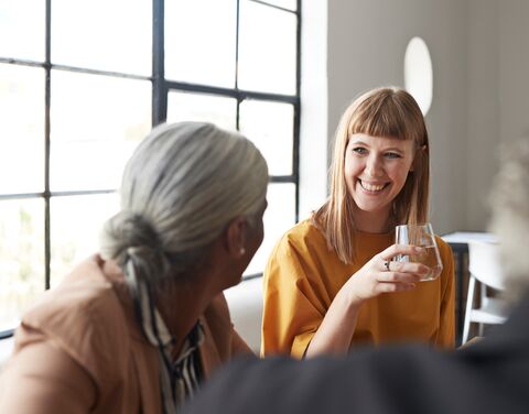 An older and a younger colleague sit in a lively conversation in a bright break room. Both laugh and gesture, indicating a friendly and engaged conversation.