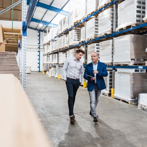 Deux hommes traversent un grand entrepôt aux hautes étagères remplies de cartons et de palettes, tout en menant une conversation et en regardant une tablette.