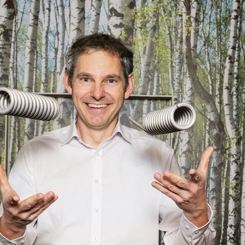 Ein Mann in einem weissen Hemd lächelt in die Kamera, während er vor einem Hintergrund mit Birkenwald steht und eine grosse Metallfeder hält.