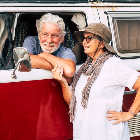 Un couple âgé sourit et s’appuie sur un camping-car rouge. L’homme regarde par la fenêtre et la femme porte un chapeau.