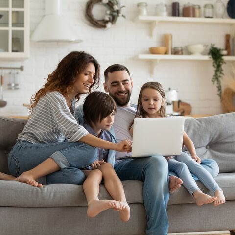 A smiling family sits together on a sofa and looks at a laptop. Everyone looks happy and is spending time together in a cozy living room.