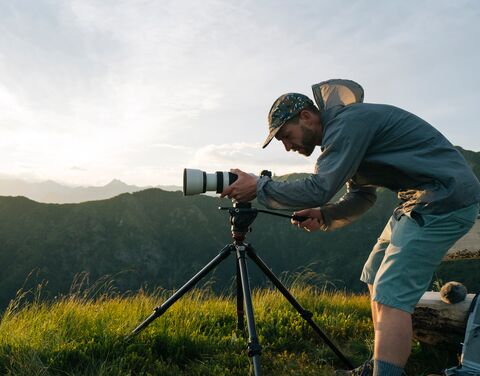 Man standing in the grass bent over his SLR camera on a tripod.