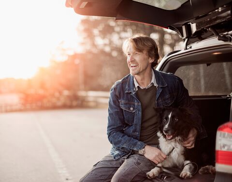 Ein Mann sitzt lächelnd mit seinem Border Collie unter der offenen Heckklappe seines Kombis, während die Sonne im Hintergrund untergeht.