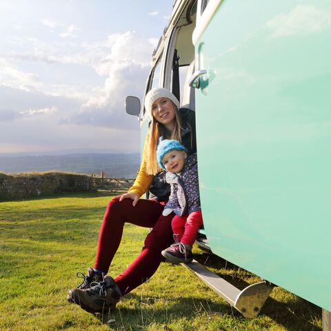 Eine Frau und ein Kind sitzen lächelnd im Eingang eines mintgrünen Campers vor einer weiten Wiesenlandschaft mit bewölktem Himmel.