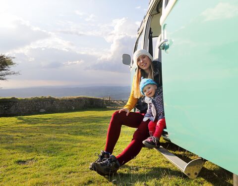 Eine Frau und ein Kind sitzen lächelnd im Eingang eines mintgrünen Campers vor einer weiten Wiesenlandschaft mit bewölktem Himmel.