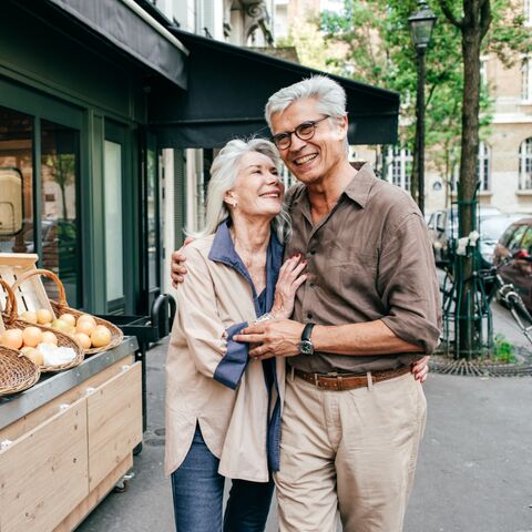 Una coppia di anziani sorride davanti a un piccolo banco di frutta all'aperto, abbracciandosi e mostrandosi felice in una giornata di sole in città.