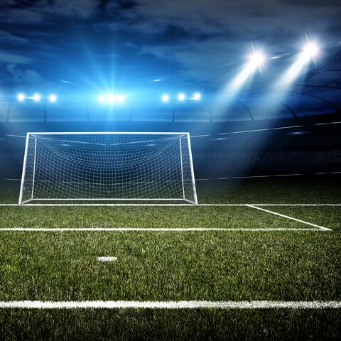 Una porta di calcio illuminata su un campo da gioco verde di notte, circondata da luci scintillanti dello stadio e da un cielo nuvoloso.