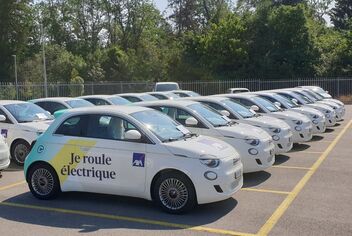 Une rangée de voitures électriques blanches sont garées sur un parking; l’une d’entre elles a un autocollant «Je roule électrique». Des arbres sont visibles en arrière-plan.