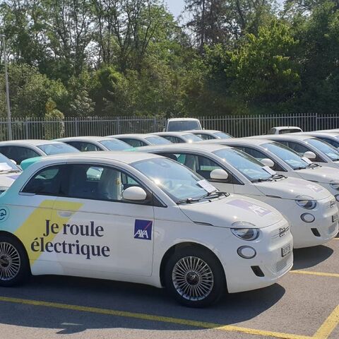 Eine Reihe weisser Elektroautos steht auf einem Parkplatz; auf einem steht «Je roule électrique». Im Hintergrund sind Bäume zu sehen.