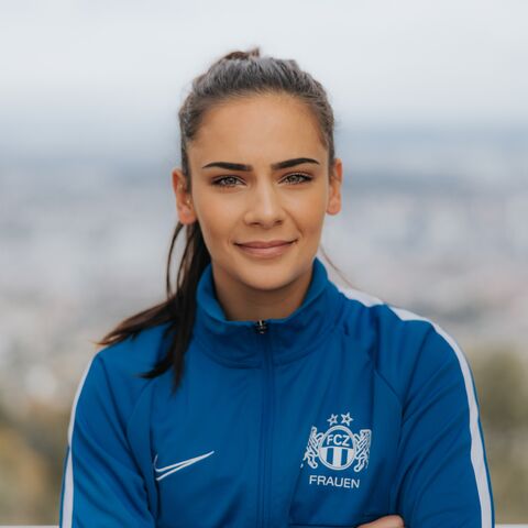 Eine Frau mit dunklem, nach hinten gebundenem Haar trägt eine blaue Sportjacke mit dem Logo des FCZ Frauen und steht draussen vor unscharfem Hintergrund.