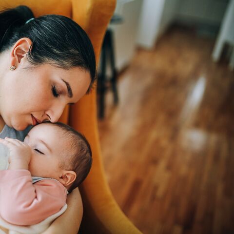 Une femme nourrit affectueusement un bébé avec un biberon et embrasse son front, assise sur une chaise dans une pièce au sol en bois.