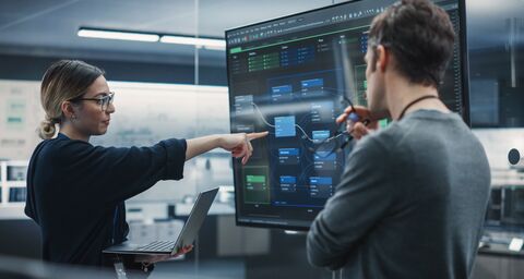 Eine Frau und ein Mann stehen vor einem grossen Bildschirm in einem Büro und besprechen etwas.