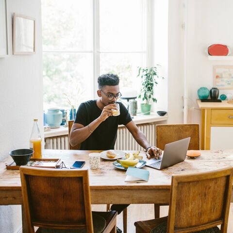 Ein Mann sitzt an einem Holztisch in einer hellen Küche, trinkt Kaffee und arbeitet an einem Laptop, umgeben von Frühstücksutensilien.