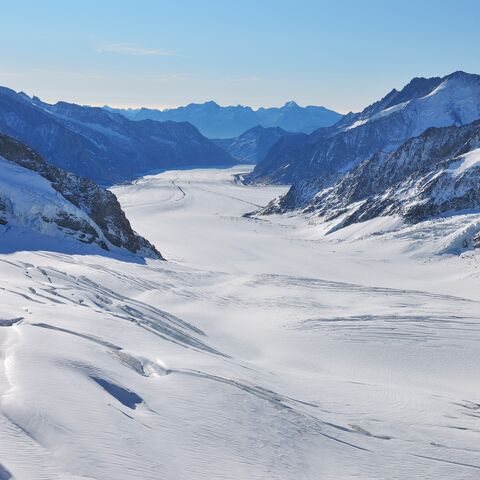 A vast, snow-covered glacier landscape with surrounding mountains under a clear, blue sky that stretches into the distance.