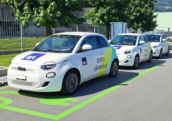 Fila di auto elettriche bianche parcheggiate sulla cui fiancata è scritto «100% elettrica». Sullo sfondo si vedono degli alberi.