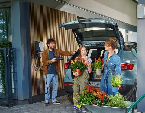 Un uomo carica un’auto elettrica mentre due donne prendono piante e fiori dal bagagliaio davanti a una casa moderna.