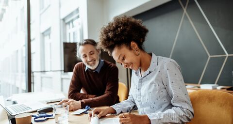 Une femme et un homme installés à un bureau travaillent en souriant. Autour d’eux, du matériel de bureau et un ordinateur portable dans un cadre moderne.