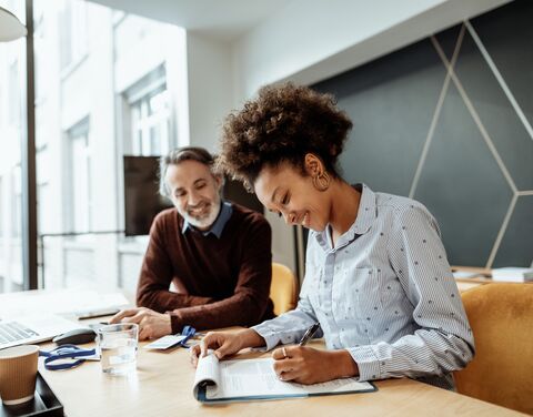 Une femme et un homme installés à un bureau travaillent en souriant. Autour d’eux, du matériel de bureau et un ordinateur portable dans un cadre moderne.