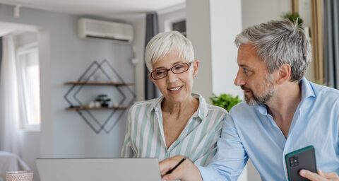 Two older people are sitting together in front of a laptop, talking about something.