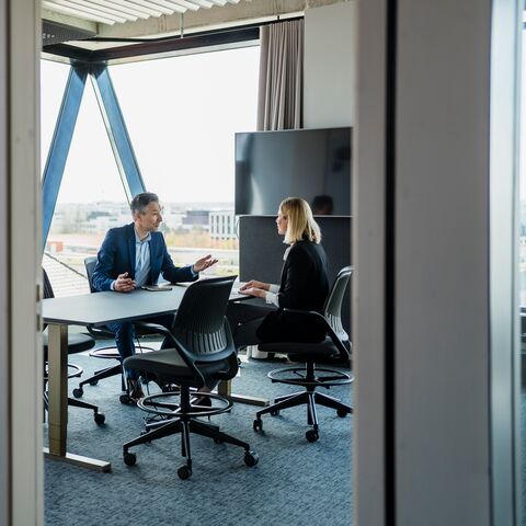 Ein Mann und eine Frau sitzen an einem Besprechungstisch in einem modernen Büro mit Glaswänden und unterhalten sich angeregt.